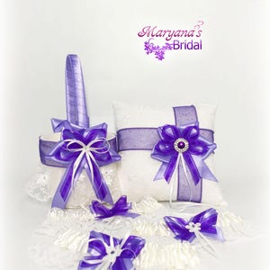 May include: A bridal set with a white lace basket and pillow, decorated with purple ribbons and bows. The set includes matching garters with purple bows. The pillow has a large purple bow with a central jewel.