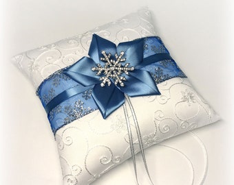 May include: A square white ring bearer pillow featuring a white fabric with silver swirling embroidery. A blue satin ribbon band wraps around the center, adorned with a blue fabric flower and a silver rhinestone snowflake. Two thin silver ribbons hang down.