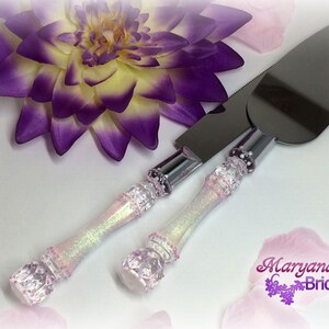 Pode incluir: Um conjunto de duas facas para servir bolo com cabos brancos iridescentes e lâminas prateadas. Os cabos são decorados com strass e têm um acabamento de glitter rosa e roxo.