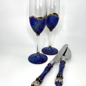 May include: Two champagne flutes and a cake serving set. The flutes and serving set have a blue, purple, and gold galaxy design. The flutes are clear glass with a gold rim. The cake server is stainless steel.