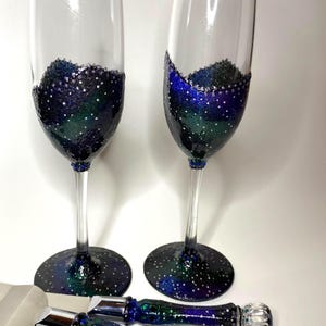 May include: Two clear glass champagne flutes and a cake serving set featuring a galaxy-inspired design. The lower bowls of the flutes, the bases, and the handles of the server and knife are decorated with deep blue, purple, and green glitter.