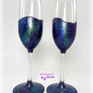 May include: A pair of champagne flutes with a celestial design. The lower half of each glass is painted with a shimmering blue, green, and purple galaxy pattern, dotted with white specks. The rims are adorned with small, clear embellishments.