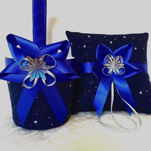 May include: A navy blue wedding basket and ring pillow set. The basket has a handle and is adorned with a large blue satin bow and a silver embellishment. The pillow is also navy blue, with a matching bow and silver accents. Both items have a starry pattern.