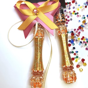 May include: A cake serving set featuring a knife and a server with handles decorated in gold glitter, orange accents, and white beads. The server handle is adorned with a pink and gold ribbon bow featuring a central pink rhinestone embellishment.