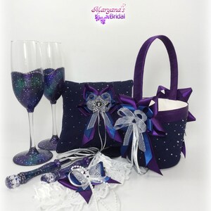 Op de afbeelding: Een verzameling bruiloftaccessoires in paars, blauw en wit. Inclusief twee champagneglazen met glitterbases, een ringkussen, een mand, een kousenband en een toverstaf. De items zijn versierd met linten en versieringen.