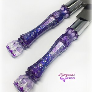 May include: A cake knife and server set featuring metallic silver blades and custom resin handles. The handles are filled with deep purple glitter, iridescent flakes, and light purple circular beads, with the text Maryana's Bridal printed.