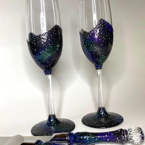Op de afbeelding: Twee champagneglazen van helder glas en een taartbestekset met een sterrenstelsel-ontwerp. De onderkant van de glazen en de handvatten van het taartmes en de taartschep zijn versierd met blauwe, paarse en groene glitters.