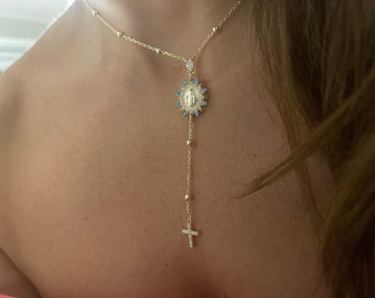 Puede incluir: Un collar de cadena dorada con un colgante que presenta una figura religiosa rodeada de piedras azules. El colgante está unido a una cadena más larga con una cruz en la parte inferior.