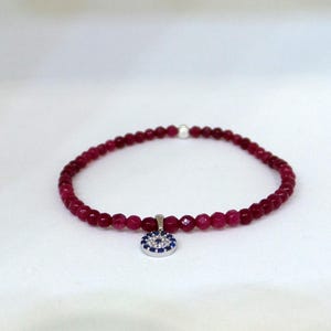 May include: A pink beaded bracelet with a silver evil eye charm. The charm has a blue eye and white accents.