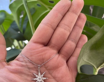 May include: A silver chain necklace with a star-shaped pendant encrusted with small, clear gemstones. The pendant has eight points and is held in a hand.