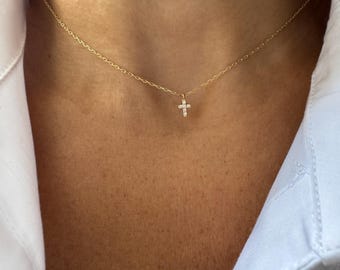 May include: A delicate gold necklace featuring a small cross pendant adorned with sparkling gemstones. The cross is centered on a fine gold chain, creating a simple yet elegant design. The necklace is shown against a skin tone, highlighting the jewelry.