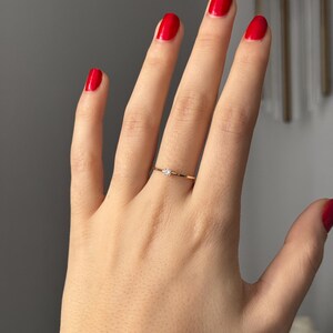 May include: A delicate gold ring with a small, clear gemstone set in the center. The ring is worn on a hand with red nail polish. The ring appears to be a minimalist design, suitable for everyday wear.