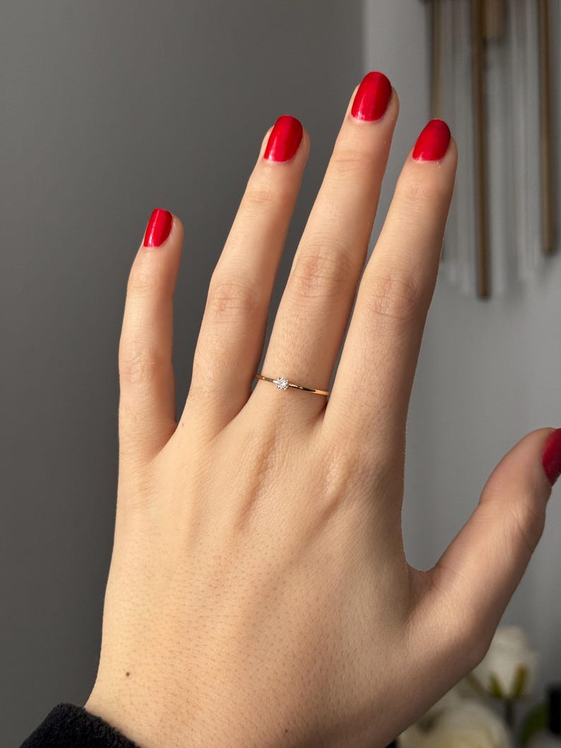 May include: A delicate gold ring with a small, clear gemstone set in the center. The ring is worn on a hand with red nail polish. The ring appears to be a minimalist design, suitable for everyday wear.