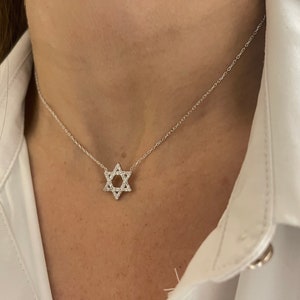 Puede incluir: Un collar de plata con un colgante de estrella de David. El colgante está engastado con pequeñas piedras brillantes y cuelga de una fina cadena. El collar se lleva sobre una camisa blanca.