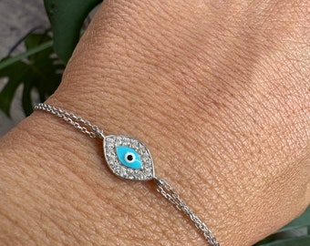 Puede incluir: Un brazalete de cadena de plata con un dije de ojo turco azul. El dije está rodeado de piedras de circonita cúbica transparentes.