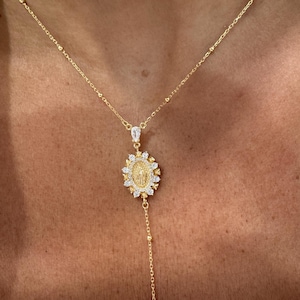 May include: Gold-toned necklace featuring a pendant with a religious figure surrounded by sparkling stones. The pendant hangs from a delicate chain with small, evenly spaced beads. A longer chain with a single bead at the end extends from the pendant.