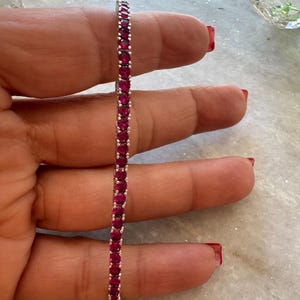 Pode incluir: Uma pulseira de ténis com uma fila contínua de pedras preciosas redondas de um rosa-avermelhado profundo, engastadas numa armação de metal prateado polido. O design consiste em pedras circulares uniformes presas por pequenas garras.