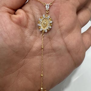 May include: Gold-toned necklace featuring a pendant with a religious design, surrounded by small, clear stones. The necklace includes a cross pendant at the bottom and a teardrop-shaped stone near the clasp. The chain is delicate and has small, round beads.