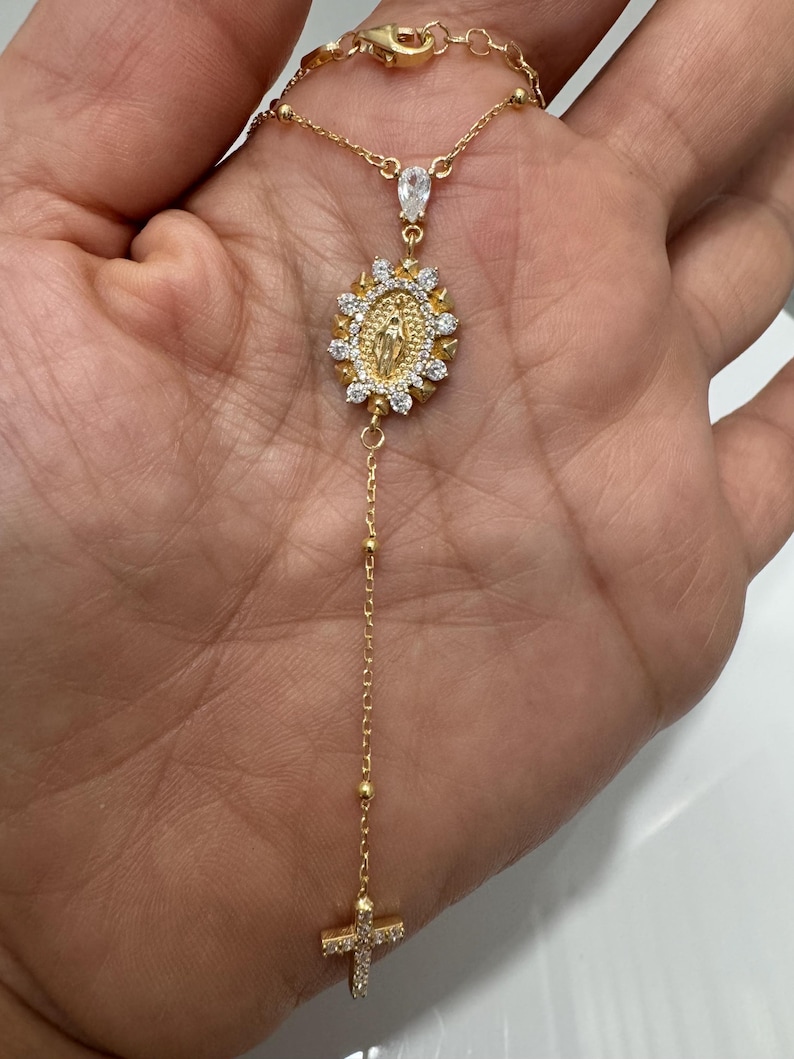 May include: Gold-toned necklace featuring a pendant with a religious design, surrounded by small, clear stones. The necklace includes a cross pendant at the bottom and a teardrop-shaped stone near the clasp. The chain is delicate and has small, round beads.