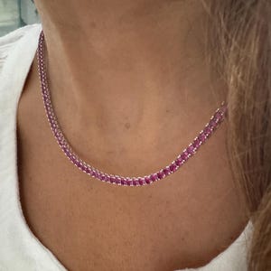 May include: A tennis necklace featuring a continuous row of round, vibrant pink gemstones set in a polished silver-toned metal chain. The necklace rests against the neck, showcasing a uniform arrangement of faceted stones that catch the light.