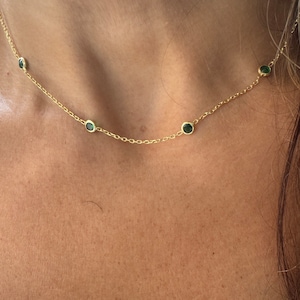 May include: A delicate gold necklace featuring small, round, emerald-green gemstones evenly spaced along the chain. The necklace is displayed on a person's skin, highlighting its simple elegance and subtle sparkle.