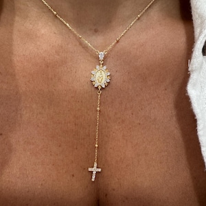 May include: Gold-toned necklace featuring a pendant with a religious motif, surrounded by sparkling stones. The necklace has a delicate chain with a cross pendant at the end. The necklace is worn against a white shirt.
