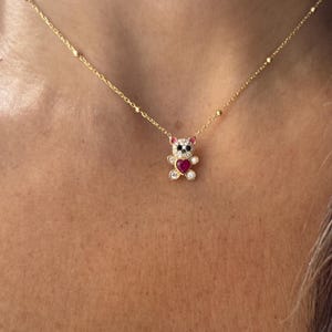 Pode incluir: Colar dourado com um pingente de urso de pelúcia. O urso tem um coração vermelho, rodeado por pedras preciosas transparentes e detalhes rosa. A corrente tem pequenas contas redondas. Uma joia delicada.