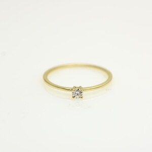 May include: A delicate gold ring with a single, small, round diamond set in the center. The ring band is thin and polished, creating a minimalist and elegant design. The ring is set against a white background.
