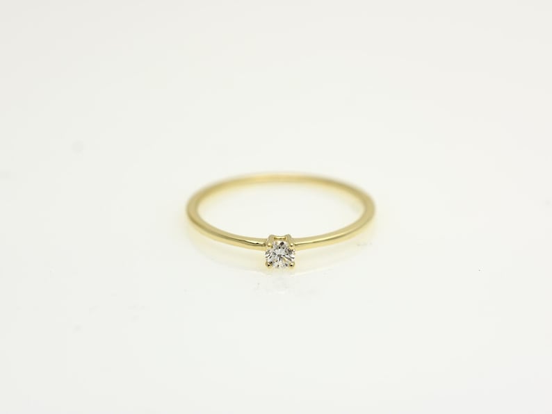 May include: A delicate gold ring with a single, small, round diamond set in the center. The ring band is thin and polished, creating a minimalist and elegant design. The ring is set against a white background.