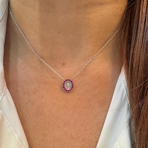 Może przedstawiać: Srebrny naszyjnik z owalną zawieszką z centralnym białym kamieniem otoczonym małymi, czerwonymi kamieniami. Zawieszka wisi na delikatnym łańcuszku. Naszyjnik jest noszony na białej koszuli.