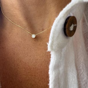 May include: A delicate gold necklace featuring a single, round, clear gemstone pendant. The necklace is displayed against a person's skin and a white button-down shirt.