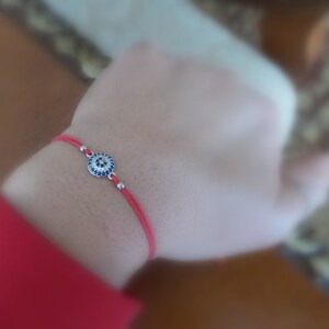 Puede incluir: Un brazalete de cuerda roja con un dije de ojo turco plateado. El dije está decorado con piedras azules y blancas.