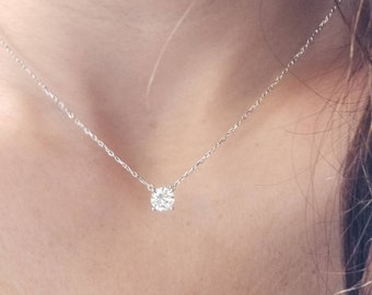 以下が含まれることがあります： 単一のラウンドカットダイヤモンドペンダントが付いた繊細なシルバーネックレス。 ダイヤモンドは、細いチェーンから吊り下げられた、クラシックな4プロングセッティングにセットされています。 ネックレスは肌の色合いの背景に対して表示されます。