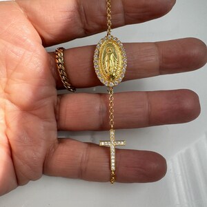 May include: A gold chain bracelet with a small cross and a pendant featuring the Virgin Mary, both adorned with clear cubic zirconia stones.