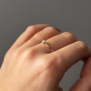 May include: A delicate gold ring with a small, clear gemstone set in a four-prong setting. The ring is simple and elegant, with a thin band. The ring is shown on a hand against a neutral background.