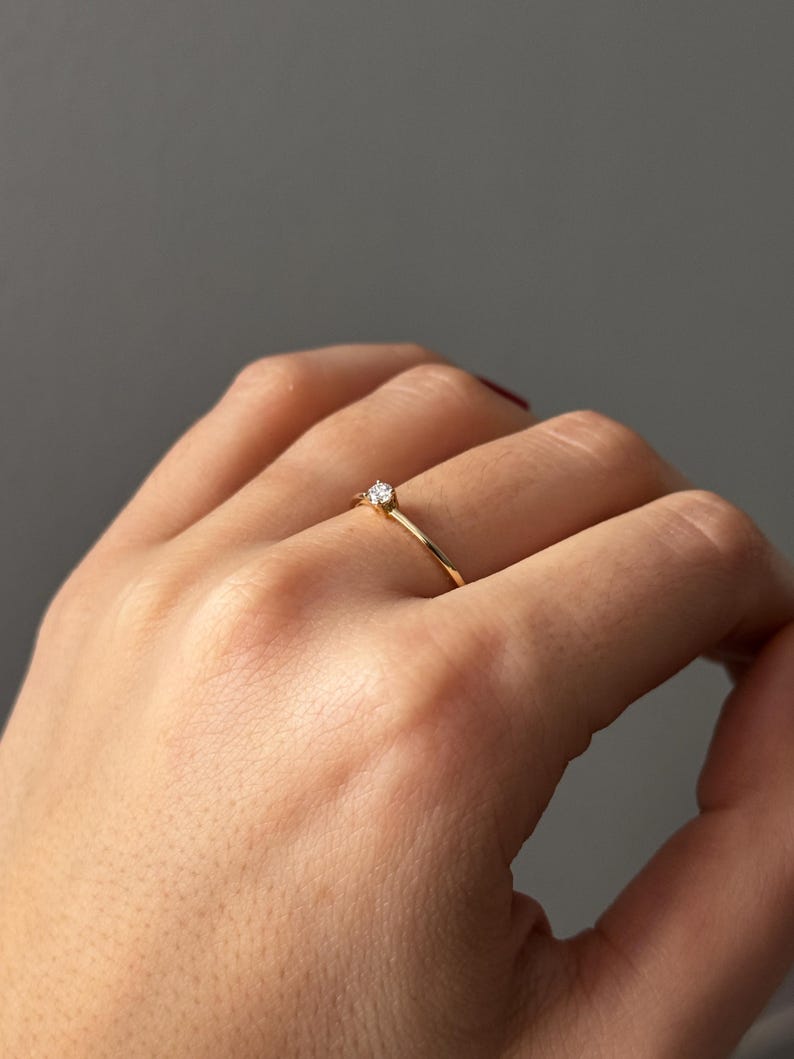 May include: A delicate gold ring with a small, clear gemstone set in a four-prong setting. The ring is simple and elegant, with a thin band. The ring is shown on a hand against a neutral background.