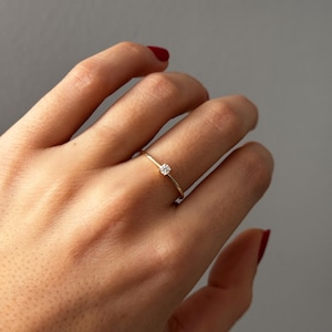 Peut inclure: Une délicate bague en or ornée d'une petite pierre précieuse transparente. La bague a un design simple, avec un fin anneau et une seule pierre étincelante. La bague est portée sur une main avec du vernis à ongles rouge.