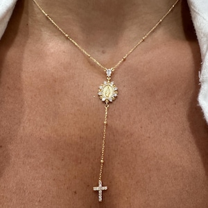 May include: Gold-toned necklace featuring a pendant with a religious motif, surrounded by sparkling accents. The necklace has a delicate chain with small beads and a cross pendant at the end. The necklace is worn against a person's skin.