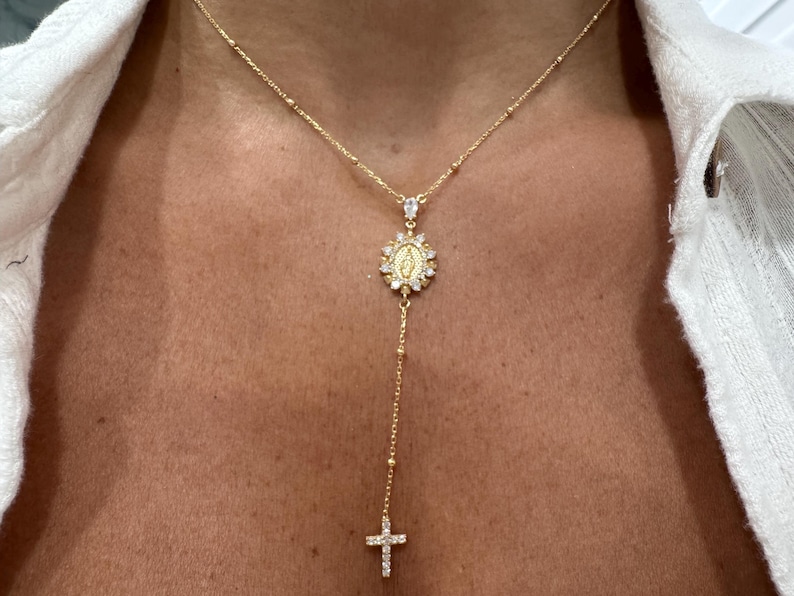 May include: Gold-toned necklace featuring a pendant with a religious motif, surrounded by sparkling accents. The necklace has a delicate chain with small beads and a cross pendant at the end. The necklace is worn against a person's skin.