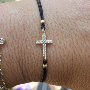 May include: A black string bracelet with a small gold cross charm set with clear cubic zirconia stones. The bracelet is worn on a wrist with other bracelets.