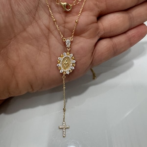 May include: A gold-colored necklace featuring a pendant with a religious motif and a dangling cross. The pendant has a central figure surrounded by a halo of white stones. The cross is also adorned with white stones. The necklace is held in a hand.