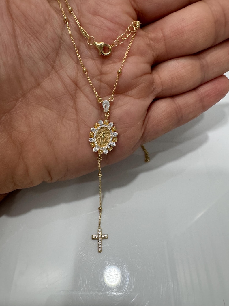 May include: A gold-colored necklace featuring a pendant with a religious motif and a dangling cross. The pendant has a central figure surrounded by a halo of white stones. The cross is also adorned with white stones. The necklace is held in a hand.