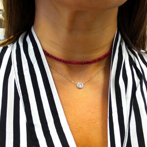 May include: A woman wearing two necklaces. The top necklace is a choker made of small, red beads. The bottom necklace is a silver chain with a single, round, clear gemstone pendant. The woman is wearing a black and white striped top.