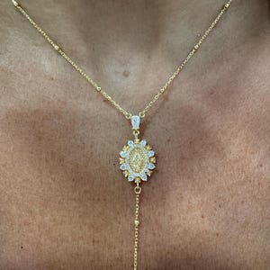 May include: A gold-colored necklace with a pendant featuring a religious figure. The pendant is oval-shaped, surrounded by small, clear stones, and hangs from a delicate chain. The necklace is worn on a person's chest.
