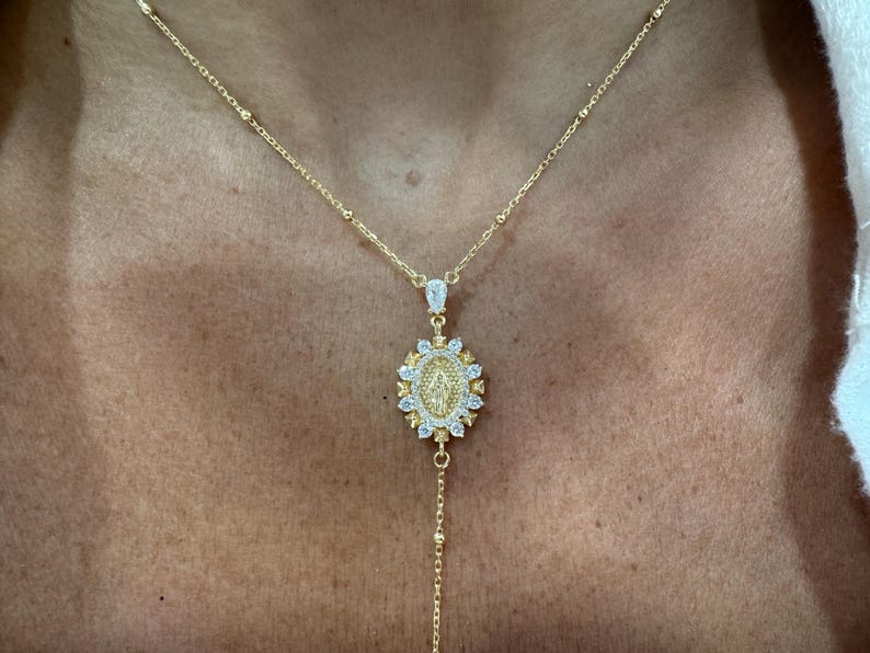 May include: A gold-colored necklace with a pendant featuring a religious figure. The pendant is oval-shaped, surrounded by small, clear stones, and hangs from a delicate chain. The necklace is worn on a person's chest.