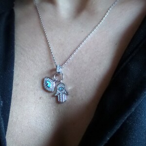 Op de afbeelding: Een zilveren ketting met een hamsa hand hanger en een boze oog hanger. Beide hangers zijn versierd met heldere strass steentjes en blauw email.
