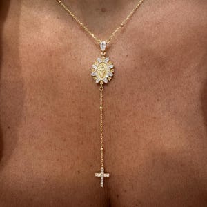 May include: Gold-toned necklace featuring a pendant with a religious motif, surrounded by sparkling accents. The necklace has a delicate chain with a cross pendant at the end. The jewelry is worn against a skin tone.