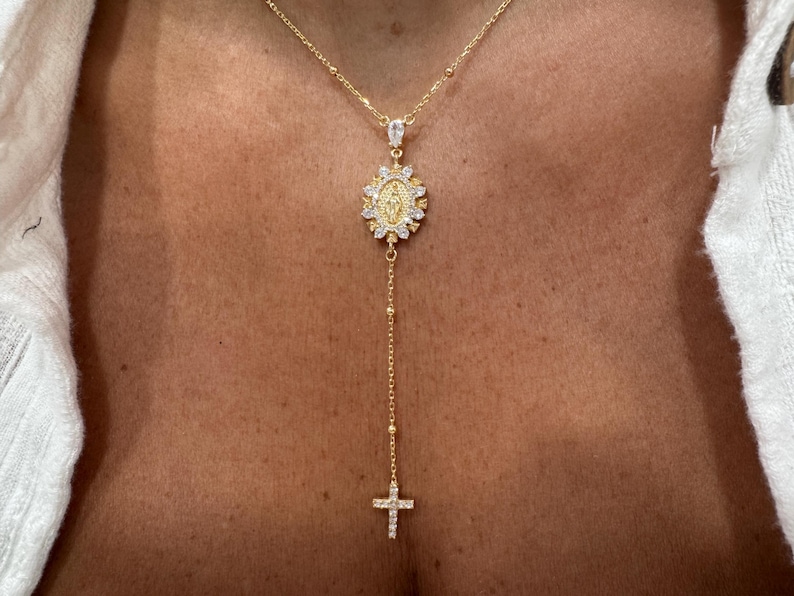 May include: Gold-toned necklace featuring a pendant with a religious motif, surrounded by sparkling accents. The necklace has a delicate chain with a cross pendant at the end. The jewelry is worn against a skin tone.
