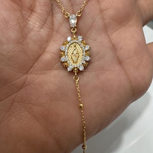 May include: Gold-toned necklace featuring a pendant with a religious figure surrounded by clear gemstones. The necklace has a delicate chain with a teardrop-shaped stone and a small cross pendant at the bottom. The necklace is displayed on a hand.