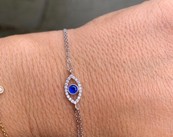 Puede incluir: Un brazalete de cadena de plata con un dije de ojo azul malvado. El dije está rodeado de pequeñas piedras transparentes.